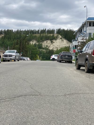 The image shows a street scene in Whitehorse, Canada, on an overcast day. A line of parked cars is visible on the right side of the street, facing away from the viewer. The cars include a silver pickup truck, a dark SUV, a white SUV, and other vehicles further down the line.
Behind the cars, a steep, sandy embankment covered in evergreen trees rises upwards. The trees create a dense forest that extends to the top of the frame. The sky is filled with heavy, gray clouds, suggesting it might be a cloudy or rainy day.
In the foreground, the asphalt road is cracked in places. To the right of the road, a modern, multi-story building with a blue roof is visible. A street sign indicating "STOP" is mounted on a pole next to the building. There are no people visible in the image. The overall mood is calm and perhaps a bit subdued due to the overcast weather.
