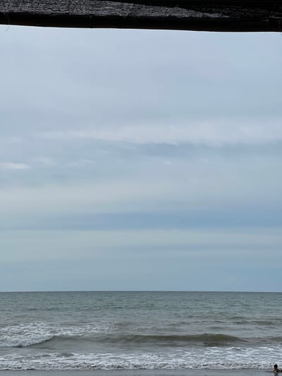 A person is lying on a wet sandy beach in front of a grey ocean under a cloudy sky. The person is partially submerged in the water. A dark mesh screen hangs from the top of the frame, obscuring the sky and a portion of the water. The scene is located on the coast of Weujangka, Indonesia.