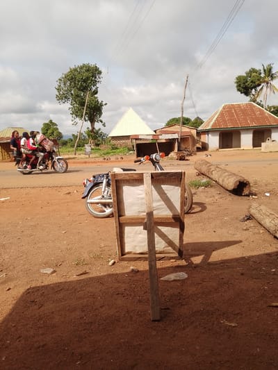 The image captures a street scene in Ichol, Nigeria. A motorcycle carrying five individuals, including a driver and four passengers, travels on a reddish-brown dirt road. The passengers are a mix of adults and children, with one passenger appearing to be a child wearing a pink top. The motorcycle is slightly angled towards the left side of the frame. In the foreground, a wooden frame with a white canvas or tarp stretches across it is positioned diagonally, with a shadow cast by it on the ground. To the right of the frame, two large logs lie on the ground. In the background, several structures with corrugated iron roofs are visible, one of which is painted white with a green trim. Trees, including a palm tree, dot the landscape. The sky is overcast with scattered clouds. Electrical wires are strung across the scene, supported by utility poles. The overall impression is of a rural or semi-urban environment in Nigeria.