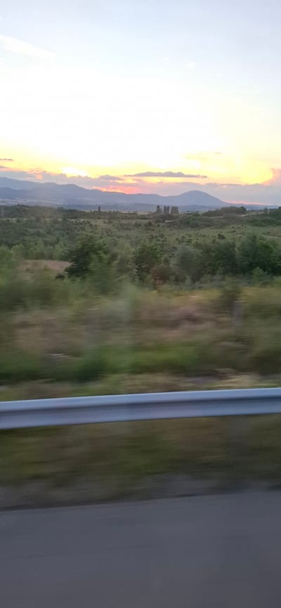 The media file is a photograph taken from a moving vehicle, likely a train or bus, looking out a window. The image is oriented vertically, giving a sense of speed and motion blur. The scene depicts a rural landscape at what appears to be sunset or sunrise.
On the left side of the frame, the sky is a bright, pale blue, transitioning into a vibrant sunset or sunrise with hues of orange, yellow, and pink closer to the horizon. Silhouetted against this colorful sky are distant mountains, with softer, lighter blue tones suggesting haze or atmospheric perspective.
The middle ground of the image features a vast expanse of greenery, indicative of fields and trees. The foliage is lush and varied in shades of green, with some patches appearing more brown or yellow, suggesting dry grass or earth. The blur of motion is evident here, with vertical streaks of color suggesting the rapid passage of scenery. A few scattered trees or small clusters of vegetation are discernible within this area.
On the right side of the frame, a distinct vertical element, likely the window frame or a part of the vehicle's interior, runs along the edge. This element is a neutral gray color and has a metallic sheen, further emphasizing the perspective from inside a moving vehicle. The blur of motion is also present in this section, but less pronounced than in the landscape.
The overall impression is one of a serene, natural landscape viewed during a transitional time of day. The combination of the soft, colorful sky and the blurred greenery creates a dynamic and somewhat dreamy atmosphere. There is no visible text in the image. The activity taking place is the observation of the landscape from a moving vehicle. The location context provided is Blatino, Bulgaria.