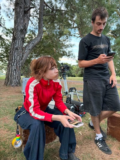 Two people are on set, likely during a break. A woman with reddish-brown hair, wearing a red jacket with a white stripe down the sleeve and dark denim pants, sits and looks at her phone. Behind her is camera equipment. A man stands behind her, also looking at his phone, wearing a black t-shirt, dark denim shorts, and black Adidas sneakers. They're outside in a grassy area with trees.
