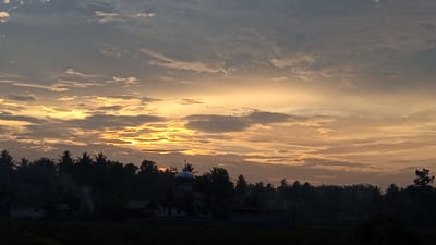 The image shows a sunset in Blangtingkeum, Indonesia. The sky is filled with clouds in shades of grey, blue, and yellow. The sun is obscured by clouds. Below the horizon, silhouetted trees and buildings are visible, including what appears to be a mosque with a dome. Smoke is emanating from behind the trees. The foreground is dark, obscuring the details of the terrain.