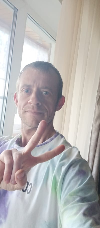 A man with short brown hair and blue eyes, wearing a tie-dye t-shirt with purple, green, and white colors, is pictured in a selfie. He is holding up two fingers in a peace sign gesture. The background shows a window with white curtains. The man is indoors, in what appears to be a residential setting in Vyazma, Russia.