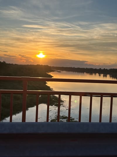 This media file captures a sunset over a body of water, possibly a river, in Diurtiuli, Russia. The scene is viewed from behind a red railing, which creates a sense of being on a balcony or bridge. The sun, a bright orb of golden light, is low on the horizon, casting a warm glow across the sky and reflecting on the water's surface. Wispy clouds streak across the sky, painted in shades of orange, yellow, and blue, indicating fair weather and a picturesque sunset.

The landscape on the opposite bank of the water is silhouetted against the vibrant sky, with the dark shapes of trees and land visible. The railing itself is composed of vertical bars and horizontal crossbars, some of which are illuminated by the setting sun. The water appears calm, with subtle ripples and reflections of the trees and sky. The overall mood is serene and tranquil, evoking a sense of peace and natural beauty. The image seems to have been taken during the golden hour, as evidenced by the warm, soft light. There is no visible text in the image.