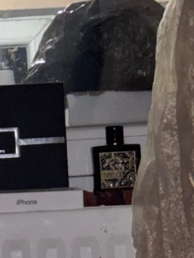 The image shows a shelf with several objects on it. In the foreground, on the right, is a section of a light-colored, textured fabric, possibly lace or a similar material. To the left of the fabric is a dark-colored perfume bottle with ornate detailing on the front. The bottle has a black cap. To the left of the perfume bottle is a black box with a silver horizontal stripe across it. Below the black box, a white iPhone box is visible with the word "iPhone" printed on it in gray. Behind the perfume bottle and black box, there appears to be a large, crumpled black plastic bag. The background is blurred but suggests a shelf or surface. The scene is indoors, and the lighting is dim. There are no people visible in the image. The location is specified as Zlataritsa, Bulgaria.