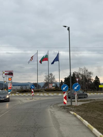 The scene depicts a multi-lane road intersection or roundabout in Dragushinovo, Bulgaria, under an overcast grey sky. In the foreground, a paved road with visible cracks and tire marks leads towards a central island. Yellow and white striped curb markings define the road edges and the central island.
On the left, a dark-colored sedan with illuminated headlights is positioned on the road, partially obscuring a Shell gas station sign featuring the company logo, a "P" for parking, and fuel prices. The gas station's building and pump canopy are partially visible behind the sign. Three flags are displayed prominently on tall poles: from left to right, a white flag with a red emblem, the national flag of Bulgaria (white, green, red horizontal stripes), and the flag of the European Union (blue with a circle of yellow stars).
Multiple blue circular traffic signs with white arrows pointing left/downwards, indicating mandatory roundabout direction, are mounted on red and white striped warning posts on the central island and the roadside. A tall, grey street light pole stands on the right side of the image, close to the road. Further back on the right, a dark grey sedan navigates the roundabout. Bare deciduous trees and evergreen trees line the area beyond the road, with rolling hills visible in the distant background. Utility lines traverse the sky horizontally above the flags. No individuals are visible.