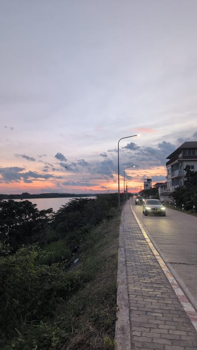 An over-the-shoulder view captures a road and sidewalk with a river and a sunset sky in Vientiane Capital, Laos. The sky displays hues of pink, orange, and gray from the setting sun, with scattered clouds. On the left, lush green vegetation lines the edge of the road, leading down to the calm river. The paved sidewalk is in the foreground, with a concrete curb separating it from the grassy embankment. The asphalt road curves to the right, illuminated by streetlights and the headlights of two moving cars. The car in the foreground is a silver sedan. Buildings are visible on the right side of the road, including a multi-story structure with balconies. The scene depicts an urban riverside setting during twilight.