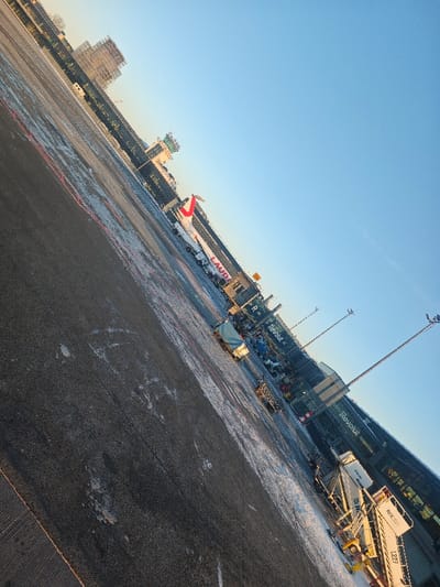 The image depicts an airport apron scene at Lidosta "Rīga", Latvia, under a clear, blue sky. The ground consists of dark asphalt with visible patches of white, indicating ice or snow. The scene is angled diagonally, with the horizon line rising from left to right.

In the mid-ground, a white aircraft with a red tail and "LAUDA" branding on its fuselage is parked, facing right, adjacent to a multi-story terminal building. The terminal structure features extensive glazing and dark facade elements, with prominent "Revolut" signage displayed on its exterior.

Numerous ground support vehicles and equipment are positioned on the apron. A set of mobile passenger stairs, marked "RIX 1221," is visible at the rear entry door of the aircraft. Additional utility vehicles, including baggage carts and a vehicle with a blue tarp, are parked near the aircraft and along the terminal facade. Two tall, metal light poles equipped with floodlights stand on the right side of the apron. In the distant background, a multi-story building under construction with visible scaffolding is present to the left of the terminal building. No specific individuals or active interactions are discernible.