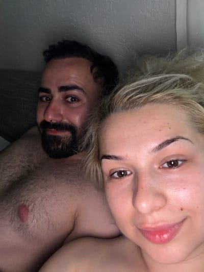 The image shows a close-up selfie of two people, a man and a woman, lying down. The woman, on the right side of the image, has blonde hair and is looking towards the camera with her eyes open. She has on light makeup, with emphasis on her eyebrows and lips. The man, on the left side, has dark, curly hair and a beard, and is also looking towards the camera. The photo is taken from a low angle, with the subjects' faces partially obscured by their hair and proximity to the camera. The setting appears to be indoors, possibly a bedroom, with a grey wall visible in the background. The lighting is soft and appears to be natural light, suggesting it could be daytime. There is no visible text in the image. The overall mood of the image seems intimate and relaxed.
