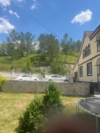 The image shows a suburban scene in Bolshoy Molokish, Moldova, on a sunny day with scattered clouds. A two-story house with a beige facade and brown trim is visible on the right side of the frame, featuring arched windows on the upper level. In front of the house, a stone wall separates the property from a street where two white sedans are parked. The background consists of a grassy, tree-covered hillside. In the foreground, a green coniferous shrub and a trampoline are situated on a grassy yard. The overall environment suggests a residential area with natural surroundings.