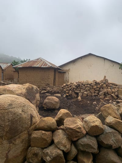 The image shows a collection of traditional mud brick houses with corrugated metal roofs in Zhwarki Mile 10, Nigeria. The houses appear to be old and weathered, with some showing signs of disrepair. In the foreground, a pile of large rocks and a wall constructed from smaller stones are visible. The environment is overcast, with a hazy sky and hints of greenery on a distant hillside. The scene conveys a sense of rural simplicity and perhaps hardship. There are no people visible in the image.