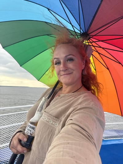 A woman with long, wavy red hair and visible makeup is holding a bottle of Corona Extra beer. She is standing on what appears to be a boat or ferry, as indicated by the railing and the water in the background. Above her, she holds a rainbow-colored umbrella, its various panels reflecting light. The woman is wearing a light-colored, collared shirt with embroidered details on the chest pocket. The sky appears overcast. The location is the city of Belous, Russia.