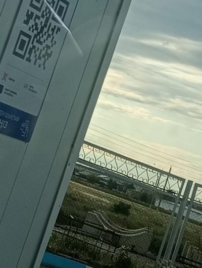 A QR code is affixed to a white vertical surface on the left side of the image, partially obscured by a transparent film. Below the QR code, a blue rectangular label displays text and a symbol. The background shows a hazy sky with clouds, and a decorative metal fence runs horizontally across the midground. Beyond the fence, a grassy area with some structures and a body of water is visible. The scene is outdoors, possibly in a public area given the QR code and fence. The location is identified as Октябрьское, Kazakhstan.