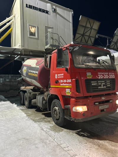 An illuminated industrial scene in Залесная, Russia, captured at night, features a red MAZ concrete mixer truck parked on a snow and ice-covered surface. The truck's cabin and mixing drum are emblazoned with "ПЕРМСКИЙ БЕТОН," "20-20-200," and "beribeton.ru." Its headlights and right-side turn signal are active, and the Russian license plate reads C245EM 159. Positioned above the truck is an elevated white industrial module, likely a concrete batching plant, bearing the "LIEBHERR" brand name. This structure includes a visible illuminated window, a door with a small access platform, and associated industrial piping, including a prominent yellow chute to the left. No individuals are present, and the scene is lit by artificial sources against a dark night sky.
