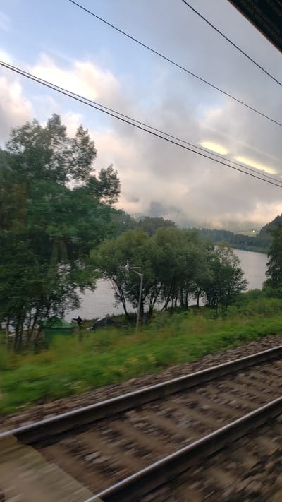 A view from a moving train shows railroad tracks in the foreground, blurred due to motion. Beyond the tracks, a body of water, likely a lake or river, is visible, bordered by a treeline and a grassy embankment. In the distance, hills and a cloudy sky are present. The scene appears to be in or around Margecany, Slovakia, a location known for its scenic landscapes and proximity to water bodies.