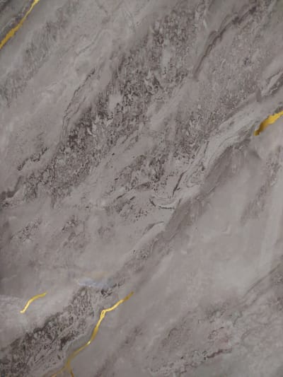 This is a close-up, high-angle shot of a textured gray surface with irregular, swirling patterns and thin, golden veins. The texture appears to be that of polished stone, possibly marble or granite, with areas of darker, more granular patches interspersed with lighter, smoother sections. The golden veins are thin and winding, adding a decorative element to the overall pattern. The lighting is even, with no distinct shadows, suggesting an indoor setting or diffused natural light. The image is a flat, abstract composition, with no discernible objects, people, or environmental context beyond the surface itself. There is no information or visual cue within the image that would allow for its location to be identified as Pitnak, Uzbekistan.