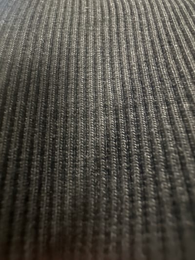 The image is a close-up, high-angle shot of a dark grey, ribbed knit fabric. The texture of the fabric, characterized by parallel vertical ridges, is the dominant feature. The lighting is uneven, creating shadows that accentuate the ribbed pattern. The background is also this fabric. There is no discernible location within the city of Letterkenny, Ireland, visible in this image, as it is a close-up of a textile.