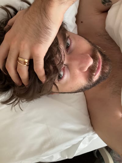 A man with dark hair, a beard, and light eyes is lying on a white pillow. His left hand is holding his hair, and he is wearing two gold rings on his ring finger. His right arm is visible behind his head, and a tattoo is visible on his right arm. The image is taken from an upside-down perspective. The location of the image is unknown, so I cannot provide the specific location in Laconia, United States of America.