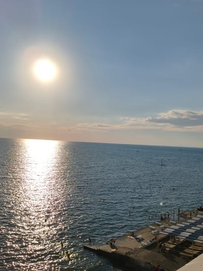 The image captures a sunny day at a beach in Blagoveschenskaya, Russia. The sun is prominently visible in the upper left quadrant, casting a bright reflection on the calm, blue sea. The reflection shimmers and sparkles, creating a dynamic visual effect. In the distance, the horizon is clearly defined, with a few scattered clouds in the light blue sky.

Along the right side of the image, a concrete pier juts into the sea. This pier is equipped with numerous lounge chairs, some covered by awnings, indicating a designated area for beachgoers. Several people are visible on the pier, some sitting on lounge chairs, while others are standing. The sea itself is populated by a number of individuals swimming or wading. A few small boats or personal watercraft are also spotted on the water. The overall atmosphere suggests a leisurely and pleasant day at the beach.