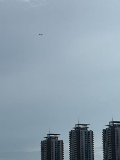 The image shows a wide shot of the sky with three tall buildings in the foreground. In the far distance, a small airplane is visible in the sky. The sky is overcast and appears to be a pale blue or grey. The buildings are modern high-rises, with glass windows and dark facades. The tallest building is on the right side of the image, with two smaller buildings to its left. The buildings are lit from above, and the sky is clear. It is not possible to determine the exact location in Pak Kret City Municipality, Thailand, from this image alone, but the presence of modern high-rise buildings suggests an urban environment.