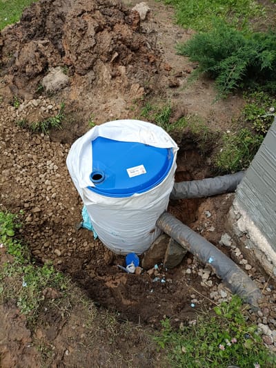 This image depicts a construction or excavation site in the city of Курапольскі сельскі Савет, Belarus. In the center of the frame, a blue plastic barrel, partially wrapped in white protective material, is situated in a dug-out hole in the earth. The barrel is connected to at least two grey flexible pipes, one of which extends towards a building's foundation visible on the right side of the image. The ground is composed of soil, gravel, and some sparse green vegetation. A pile of excavated dirt is visible in the upper left quadrant of the image, and a small green bush is present in the upper right. The scene appears to be outdoors, with natural light present. There are no people or animals visible in the image.