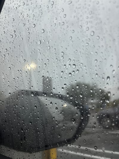 The image depicts raindrops on a car window, viewed from inside the vehicle. The background is blurred, showing a street or parking lot scene with trees, a street light, and other vehicles. The weather appears overcast and rainy, creating a somber and reflective mood. The location is identified as Tangelo Park, United States of America.