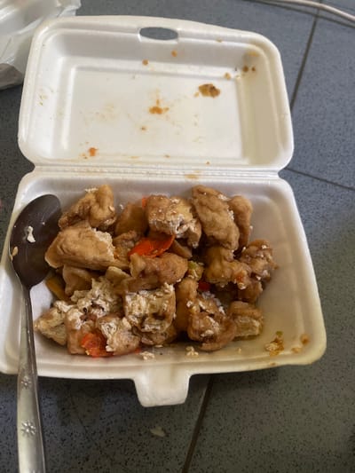 A close-up overhead shot shows a white styrofoam takeout container filled with fried tofu pieces and some red chili peppers. A silver spoon rests on the left side of the container, partially submerged in the food. The container is open, with the lid resting behind it. The food appears to be slightly messy, with some pieces broken and scattered. The background consists of a dark gray tiled surface. There are some faint orange stains on the inside of the lid of the container, indicating previous food residue. A few strands of dark hair are visible on the tiled surface near the container. The scene suggests a casual meal setting, likely in a home or outdoor environment, in Anggaswangi, Indonesia.
