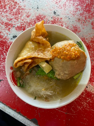 A close-up, overhead shot captures a bowl of bakso, a traditional Indonesian meatball soup, placed on a distressed red table. The bowl, white with a red rim featuring the word "SEASONING" in red text, contains a large, pale meatball topped with a vibrant orange chili paste. Adjacent to the meatball are several golden-fried wontons, layered and crinkled. Below these elements, a tangle of translucent noodles peeks through the clear, yellowish broth. Scattered amongst the noodles are green leafy vegetables, possibly bok choy, and small pieces of minced meat. The background is dominated by the textured red surface of the table, marked with white speckles and scratches, suggesting wear and tear. The scene is devoid of any people or explicit action, focusing solely on the presentation of the food. The location is identified as Sungai Sipai, Indonesia, though no specific landmarks are visible in the image to confirm this.