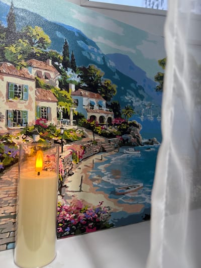 A close-up, slightly angled shot shows a canvas painting of a Mediterranean coastal town. The painting depicts white buildings with red-tiled roofs, some with blue shutters, nestled on a hillside overlooking a turquoise bay. Lush green trees and vibrant pink flowers are painted throughout the scene, and several boats are visible in the water. In the foreground, to the left of the painting, is a tall, cylindrical candle in a clear glass holder. The candle is cream-colored and emits a warm, orange artificial flame. A sheer white curtain is partially visible on the right side of the image, obscuring some of the painting and the background, which appears to be a window. The overall lighting suggests an indoor setting, possibly near a window. There are no people or animals present in the image. The setting is a still life arrangement.
