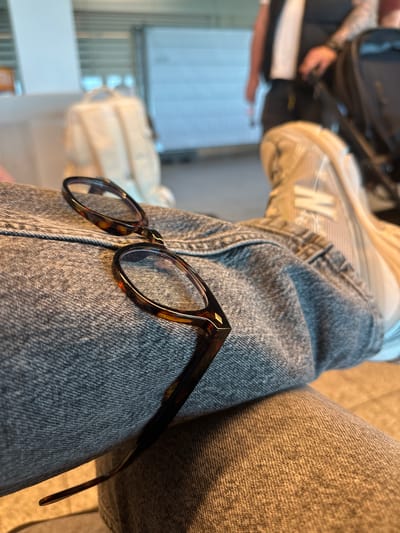 The image presents a close-up, angled perspective focusing on a pair of tortoiseshell eyeglasses resting on a person's leg, which is clad in textured grey denim jeans. The eyeglasses have a classic, rounded frame with a distinctive mottled brown and amber pattern.
In the upper left portion of the frame, the top of a light-colored sneaker is visible on the same leg, featuring a prominent white "N" logo, indicative of a New Balance shoe. The background is softly blurred but reveals elements typical of an indoor public waiting area. To the left, a shadowy figure, likely another person, is partially visible with what appears to be a dark backpack and an arm displaying a tattoo. To the right, a light-colored, possibly white, piece of luggage or a bag is indistinctly seen.
The setting appears to be a spacious indoor environment with bright, indirect lighting, suggesting a daytime scene, possibly within an airport or a large transport terminal, consistent with the location context of Weeze, Germany. The overall atmosphere is relaxed, with the subject seemingly resting or waiting. No specific activity beyond sitting and waiting is clearly depicted. No other explicit text is visible besides the "N" logo.