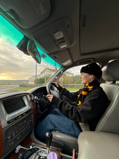 The image shows the interior of a right-hand drive vehicle, appearing to be a Toyota Land Cruiser, with a person seated in the driver's seat. The driver, wearing a black beanie, a black long-sleeved top with thumbholes, blue jeans, and a black and yellow striped scarf, holds the steering wheel with both hands and looks towards the right. The dashboard features wood-grain trim, a central display indicating "7°C" and "95.3," and an automatic gear shifter. A white charging cable is plugged into the console, where a purple container and a pink object are also present. The vehicle's windshield has a green tint at the top.

Through the windshield, a light-colored building with a sign prominently displaying "Winds Ridge Cafe" is visible across a paved area. This establishment is located at Ferry Road, Saltash, United Kingdom. Beyond the cafe, a low stone wall lines the roadside, and scattered trees with sparse foliage suggest a cooler season. In the distant left background, a red vehicle and additional red structures are partially visible, possibly train carriages or industrial elements. The sky above is overcast, with brighter patches of light.