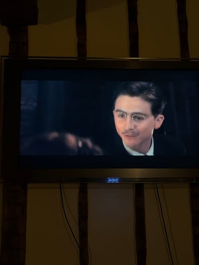 An indoor scene in Mauléon-d'Armagnac, France, depicts a dark-framed Sony television displaying a film or program. The screen features a male subject from the chest up, positioned slightly right of center. The man has dark, neatly styled hair, wears small, round, wire-rimmed spectacles, and a light mustache. He is dressed in a white collared shirt beneath a dark jacket or suit, and his mouth is slightly open, suggesting speech or an upcoming utterance. A blurry, out-of-focus human figure or object is partially visible to the left on the screen. The television is situated against a wall characterized by prominent dark wooden beams arranged vertically and horizontally, contrasting with the lighter, possibly cream-colored, plastered sections. Two dark cables descend vertically from behind the television along the wall. The ambient lighting in the room is dim, with the television screen being the dominant light source for the depicted content.