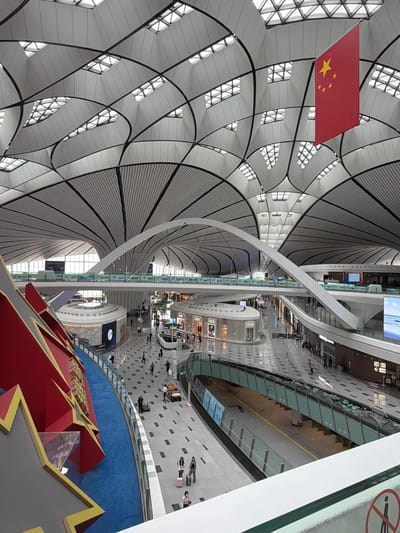 This is a wide-angle interior shot of the Beijing Daxing International Airport in Guangyang District, China. The airport's interior features a striking, modern architectural design with a vaulted ceiling composed of interconnected white panels and a grid of skylights. A large Chinese flag hangs prominently from the ceiling.

In the foreground, a large red and gold decorative structure, possibly a sculpture or part of a display, curves into the frame on the left. A blue carpeted walkway runs alongside this structure.

The mid-ground reveals a busy airport concourse with multiple levels and walkways. Shops, including what appears to be a Dior store, are visible on the lower level. People are seen walking, some with luggage, suggesting ongoing travel activity. Escalators and bridges connect different sections of the airport.

The background shows more of the airport's vast interior, with large windows or glass facades allowing natural light to enter. Digital displays and signage are visible, indicating airport services and wayfinding. The overall environment is clean, spacious, and well-lit, indicative of a major international transportation hub. The scene is captured from an elevated vantage point, looking down into the concourse.