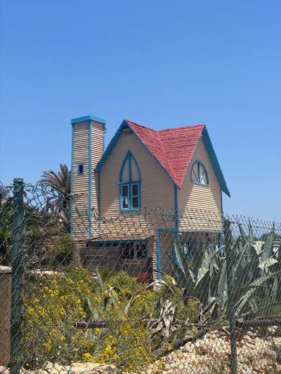 The image shows a whimsical, brightly colored house with a red roof and light yellow siding, accented with blue trim. The house features unique architectural elements, including a distinctive triangular window with blue framing and a smaller rectangular window on a protruding section. The structure is partially obscured by a chain-link fence and lush green foliage, including large agave plants and flowering yellow shrubs. The background is a clear, bright blue sky, suggesting a sunny day. The perspective is from below, looking up at the house, which is tilted on its side in the image. The overall scene evokes a sense of a charming, perhaps slightly surreal, building in a Mediterranean or arid climate. There is no visible text in the image. The location is identified as Mellieħa, Malta.