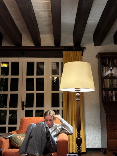 The scene depicts the interior of a room in Huasca de Ocampo, Mexico, observed at night. A young female individual is seated in a prominent orange upholstered armchair, positioned towards the left-center of the frame. She wears a light grey long-sleeved sweater and dark, vertically striped wide-leg pants. Her light brown hair is pulled back, and she is leaning forward, looking downwards, with her left hand lightly touching her temple. A green throw pillow with an embroidered design rests against her back in the armchair. To her left, a dark-covered book or tablet is visible on a surface next to the chair.
The room features a rustic aesthetic with a white textured plaster or stucco wall on the right and a ceiling characterized by dark exposed wooden beams contrasting with white plaster. A tall, dark wooden floor lamp with a cream-colored tapered lampshade, emitting warm light, stands to the right of the seated individual. Behind the armchair, large white-framed French doors or windows with multiple panes look out onto a dark exterior, showing faint reflections of interior lights. Yellow pleated curtains are drawn partially to the left of these doors. On the far right, a dark wooden hutch or bookshelf filled with books is partially visible against the textured wall. The floor in the foreground shows a few small, dark, possibly decorative objects.
