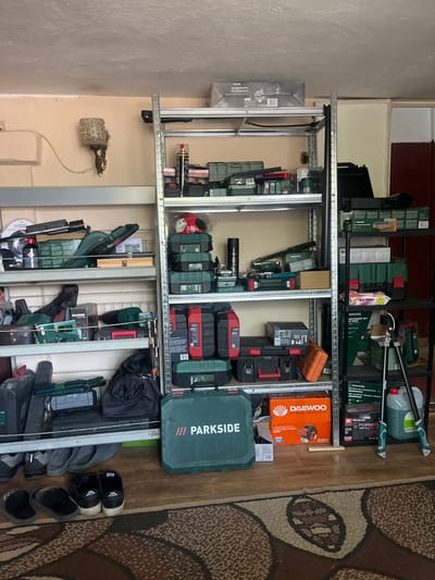This media file shows a storage area in what appears to be a garage or workshop. The scene is filled with various tools and equipment, organized on metal shelving units. Prominently displayed are power tools like drills, saws, and what looks like an air compressor, along with their associated cases and accessories. Several items are labeled with brand names such as "PARKSIDE," "DAEWOO," "RAIDER," and "Einhell." Other objects include toolboxes, storage containers, and even a few pairs of shoes on the floor. A distinctive patterned rug is visible on the right side of the image. The lighting suggests an indoor setting, likely during the daytime. There are no people visible in the frame. The overall impression is one of organized, functional storage for a collection of tools. The visible text includes brand names and product descriptions, such as "Wet Towel," "Compressed Air Reel," and "ENGINE PACK." The location context suggests this storage area is in Sadovo, Bulgaria.