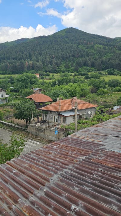 A high-angle, outdoor shot depicts a scene in Hristo Danovo, Bulgaria. The foreground is dominated by a corrugated metal roof, heavily rusted and textured. Buildings with orange tile roofs are nestled amidst dense green trees and vegetation in the midground, set against a backdrop of a steep, forested hillside rising to the left. The sky above the hillside is blue with scattered white clouds. A road cuts through the village near the buildings. A street sign stands in front of one of the buildings.