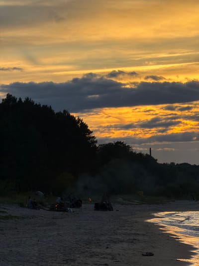 The image depicts a sunset scene at a beach in Plieņciems, Latvia. The sky is filled with dramatic clouds, illuminated by the setting sun in vibrant hues of yellow and orange. A dark, silhouetted treeline runs along the horizon, separating the sky from the beach. On the beach, a group of people are gathered around a bonfire, casting a warm glow. Some individuals are seated, while others appear to be standing or interacting with the fire. The waves of the Baltic Sea are visible on the right side of the image, with their edges reflecting the warm light of the sunset. The overall mood is peaceful and convivial, suggesting a gathering of friends or family enjoying a summer evening by the sea. The time of day is clearly sunset, and the weather appears to be clear and calm. There is no visible text in the image.