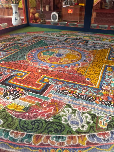 The image depicts a close-up, low-angle view of a sand mandala, a Tibetan Buddhist tradition. The mandala is intricately designed with vibrant colors and geometric patterns, crafted from finely colored sand. Its surface appears textured and granular. The mandala is enclosed within a clear glass casing, suggesting it is a display piece, possibly in a museum or temple. Reflections of the surrounding environment are visible on the glass, including a white vase with blue patterns, a stack of red apples, and a brass lamp. In the background, through the glass, there are hints of an indoor space with wooden furniture, a red sign, and a circular electronic device. The environment suggests a cultural or religious setting, likely a temple or a cultural exhibition space in Việt Trì, Vietnam, where sand mandalas are sometimes created and displayed.