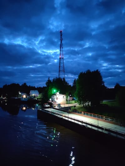 The image depicts a nighttime view of Annensky Most, Russia. A prominent communication tower stands tall, emitting red lights at its top. Below the tower is a pier or dock extending into a body of water, with white lights illuminating its surface. A small building with a green light is situated on the pier. Across the water, there are houses with lights on, their reflections visible in the water. The sky is filled with dark, cloud-filled clouds. Life preservers are attached to the railing of the pier.