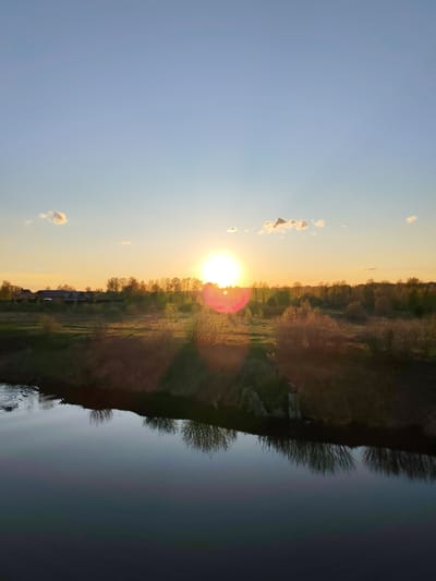 The image shows a rural landscape at sunset, possibly in Myshkin, Russia. A calm body of water, likely a river or canal, reflects the vegetation along its banks. The opposite bank is covered in trees and shrubbery, with hints of buildings or structures in the distance. The sun is setting, creating a bright glare and a warm glow over the scene, with some clouds in the upper portion of the sky. The foreground is out of focus.