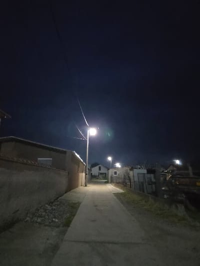 The image depicts a nighttime scene of a narrow, concrete or asphalt path in Negorci, North Macedonia, extending into the background. The sky is dark, clear, and featureless, displaying a deep blue-black hue.

On the left side of the path, a low, light-tan concrete or stucco wall runs parallel to the foreground. Behind this wall, a building with a light-colored facade and a dark roofline is partially visible, featuring a rectangular window. A vertical pole supporting a bright white streetlight stands directly adjacent to the left edge of the path, emitting strong illumination and a visible lens flare. Further down the path on the left, additional structures are vaguely discernible in the dim light.

On the right side of the path, a strip of sparse, dry vegetation and exposed soil separates the path from a rough, low fence or barrier. Beyond this barrier, a cluttered area contains multiple indistinct structures, possibly sheds or utility buildings, along with accumulated materials or debris. Another bright streetlight is visible further down the path on this side.

Multiple bright, white streetlights provide the primary illumination for the scene, casting light onto the path and adjacent structures. Thin black wires, likely power or communication lines, are visible against the dark sky in the upper portion of the image. No individuals, animals, or vehicles are present in the depicted scene.