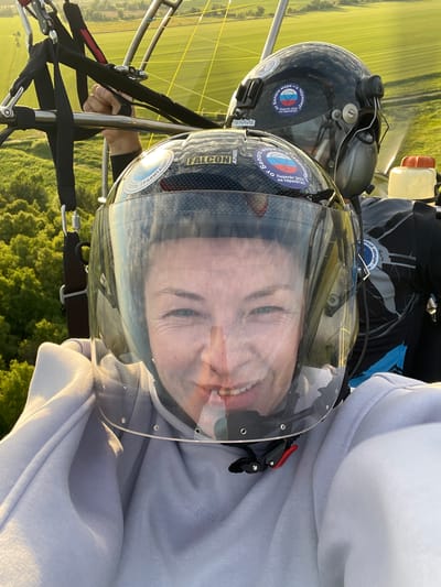 The image shows a selfie taken from a paraglider piloted by two individuals. In the foreground, a woman with a clear visor helmet smiles. The helmet has a sticker with a Russian flag and text. Behind her, another person in a black helmet is visible. The paraglider is equipped with metal bars and straps. The background consists of green fields and trees, suggesting a rural or suburban environment. The context implies the location is in or near the village of Турово, Russia, given the metadata.