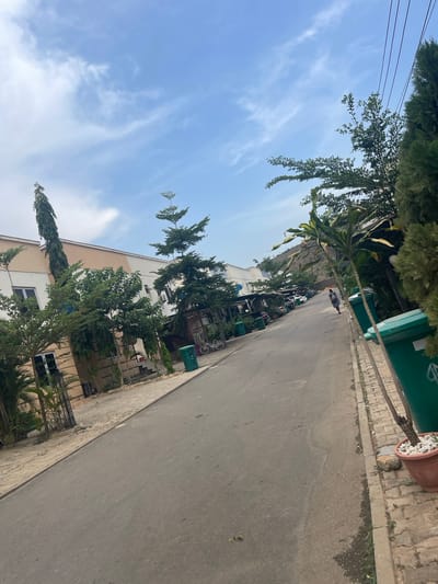 This media file captures a daylight scene of a street or pathway in Tasha, Nigeria, featuring buildings, lush greenery, and a clear sky.
The central subject is a paved asphalt road, extending into the distance. Parallel to it on the right is a brick-paved sidewalk, where a green trash bin and a large potted plant filled with white decorative stones are visible. Trees and various plants densely line both sides of the road, creating a verdant corridor. Some trees have broad canopies, while others are tall and slender, or feature hanging foliage. Power lines are visible across the upper left portion of the sky.
Behind the vegetation, multi-story buildings, likely residential or commercial, appear on both sides. These structures are predominantly light-colored (beige and white) and feature multiple windows. In the mid-ground on the left side, behind the trees, some parked vehicles are partially visible, along with bicycles leaning near buildings.
A single person is captured in the mid-ground, walking away from the camera on the right side of the road, dressed in light clothing. The activity depicted is ordinary street-level movement, suggesting a calm, everyday setting.
Notable details include a bright blue sky with wispy white clouds, indicating clear weather and good visibility. The time of day appears to be mid-morning or early afternoon due to the strong, even lighting. While there is a logo or text on the green trash bin, it is not legible. The overall impression is one of a functional, tree-lined street in an urban or suburban environment.
