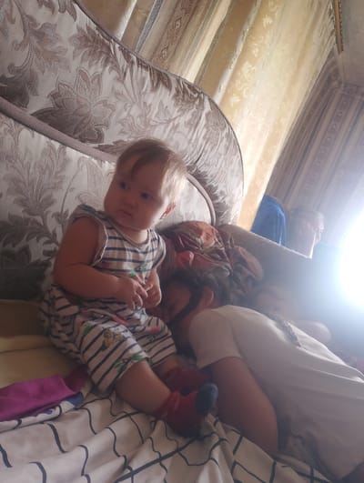 A baby wearing a striped onesie and red socks sits on a bed, looking to the left. Behind the baby, two adults are lying down, partially obscured by the baby and the bright sunlight. The room appears to be a bedroom, with a patterned headboard and draped curtains visible in the background. The bed linens have a black and white striped pattern. The overall impression is one of a casual, home environment. The location of Лобаново, Kazakhstan is noted.