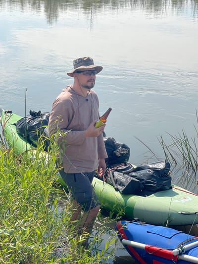 A man wearing a camouflage bucket hat, sunglasses, a tan hooded shirt, and gray shorts stands in shallow water next to a green inflatable raft. The raft is loaded with several large black trash bags. A blue inflatable raft is partially visible in the foreground, in front of the green raft. The man is holding a brown glass bottle in his right hand. He is looking to his right. The water appears calm with reflections of trees on the surface. The scene is outdoors with green vegetation on the left side of the image. The location is identified as Nezhinka, Russia.