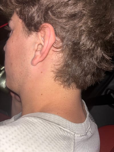 The image displays a close-up, profile view of the right side of an individual's head, neck, and upper torso. The subject is an adult male with fair skin, prominent right ear, and dark brown, wavy hair that extends to the nape of the neck. Stubble is visible along the jawline. The individual is wearing a light grey, textured, long-sleeved shirt, possibly a thermal knit, with visible stitching details on the right shoulder. The background is predominantly dark, suggesting an indoor or low-light environment. A faint light source or reflection is present on the far left, and a portion of a red surface, possibly upholstery or fabric, is visible on the far right below the shoulder. The specific location within the city of Quincy, United States, cannot be determined from the visual information in the image.