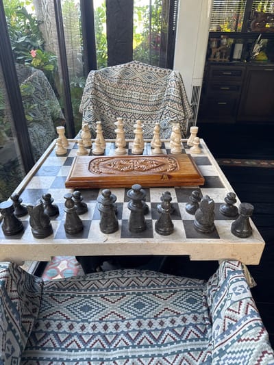 The scene depicts an unoccupied chess game setup inside a room in Tibubiyu, Indonesia, illuminated by natural daylight. A square chess board, featuring alternating light and dark grey marble or stone squares, rests on a light-colored stone table or stand in the foreground.
On one side of the board, a complete set of cream-colored chess pieces, appearing to be made of carved bone or a similar light material, are arranged in their initial positions. Centered on the first two ranks of this side, replacing some initial pieces, is an oblong, carved wooden box with intricate scrollwork designs.
Opposite these, a complete set of dark grey, stone-like chess pieces are arranged in their initial positions on the corresponding ranks. The white king is on E8, queen on D8; the black king is on E1, queen on D1 (relative to white's setup).
Behind the cream-colored pieces, a chair is draped with a patterned fabric displaying geometric diamond and zigzag motifs in cream, light blue-grey, and reddish-brown. Large, dark-framed glass panels or windows form the back wall, revealing lush green outdoor foliage and trees, with reflections visible on the glass.
To the right, a tall white appliance, likely an air conditioner, stands beside dark wooden cabinetry. The floor in this area is dark. Beneath the chess board, the top portion of a second chair or seat is visible, covered in a similar patterned fabric, along with a section of a floor or rug with colorful geometric patterns in red, blue, and white. No individuals are present in the image.