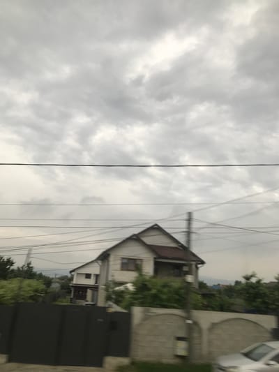 The image captures a residential street scene in Nicolae Bălcescu, Romania, under a heavily overcast sky dominated by dense grey clouds. Multiple black power lines traverse horizontally across the mid-ground, with a prominent thicker line at the top and several thinner lines below, partially obscuring the distant background. A two-story house with a white facade and a dark brown pitched roof is centrally positioned on the right, featuring a visible upper-story window and a recessed lower-story balcony or porch. An additional white structure with a darker roof is partially visible to the left of the main house. Lush green vegetation, including trees and bushes, surrounds the properties and extends into the background. In the foreground, a dark grey, solid fence or gate occupies the lower-left, while a light grey concrete wall with decorative arched sections is present on the lower-right. A utility pole is visible centrally, intersecting the power lines and the concrete wall. The blurred front section of a silver car is evident in the lower-right corner, indicating motion at the time of capture. Distant, faint hills are discernible on the horizon beneath the cloudy sky. No individuals are visible.