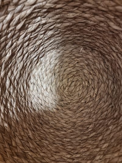The image is a close-up, overhead view of the interior of a woven basket. The basket is made of natural fibers, likely straw or raffia, and exhibits a tight, concentric weave pattern that spirals towards the center. The texture is rough and fibrous, with variations in color from light tan to darker brown, suggesting natural material. There are no people, animals, or visible actions taking place in the image. The lighting is natural and somewhat diffused, highlighting the intricate details of the weave. There is no identifiable location in the city of Pohranov, Czechia.